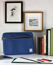 Load image into Gallery viewer, A 15 inch MacBook case in blue shade shows the patterned lining inside the pocket as it sits on a desk next to a row of sketchbooks and design manuals.
