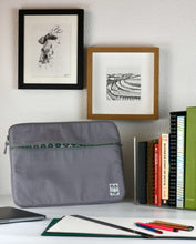 이미지를 갤러리 뷰어에 로드 , A grey 15 inch laptop case sits on a white table with pencils and a sketchpad ready for use. On the white wall behind are some pen and ink drawings of the shells on the sea shore and a snowy field with tractor markings.
